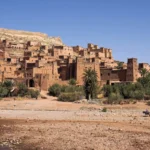 The earthen ksar of Aït-Ben-Haddou rising in tiers against a hill above palm groves and a dry riverbed in Morocco