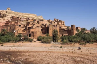 The earthen ksar of Aït-Ben-Haddou rising in tiers against a hill above palm groves and a dry riverbed in Morocco