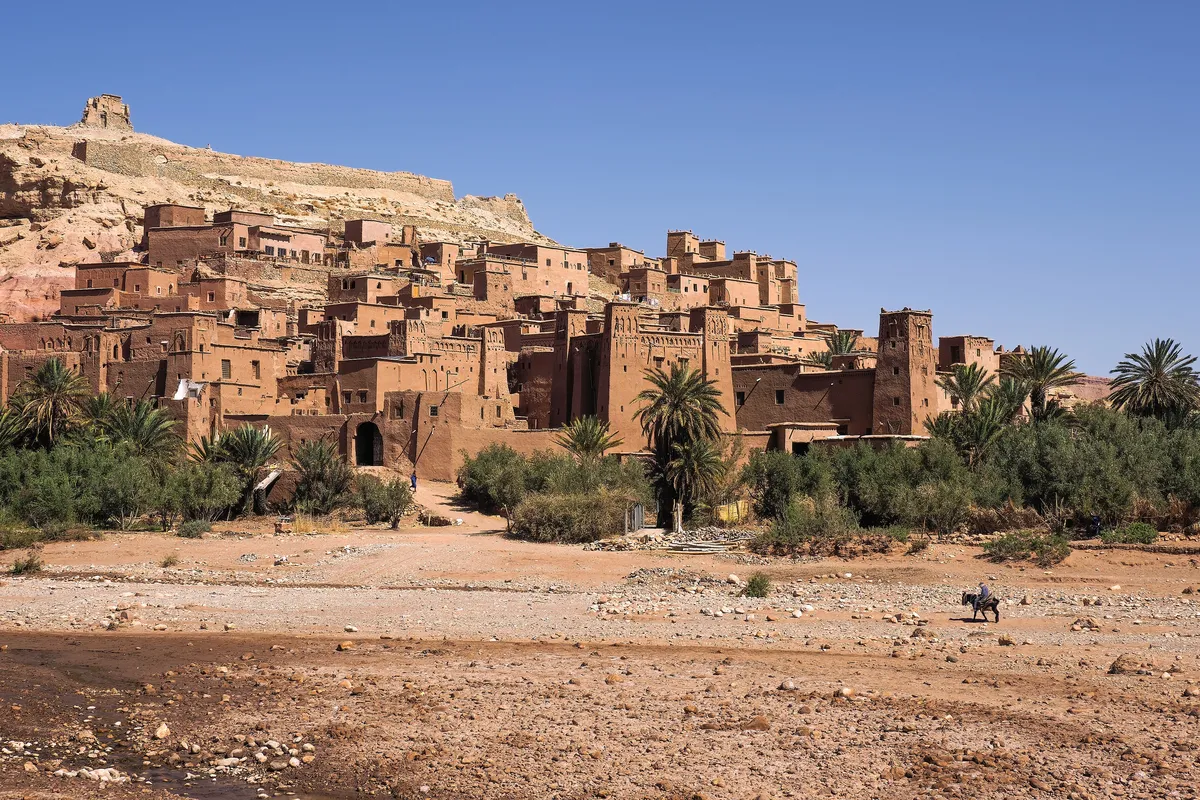 The earthen ksar of Aït-Ben-Haddou rising in tiers against a hill above palm groves and a dry riverbed in Morocco